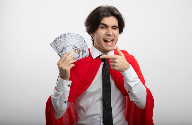 Joyeux jeune homme de super-héros portant une cravate tenant et des points à l'argent isolé sur blanc