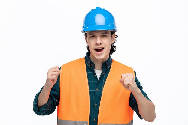 Joyeux jeune ingénieur masculin portant un casque de sécurité et un gilet de sécurité regardant la caméra faisant un geste oui isolé sur fond blanc