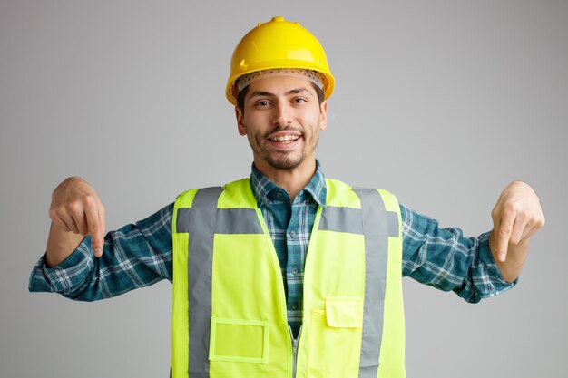 Joyeux jeune ingénieur masculin portant un casque de sécurité et un uniforme regardant la caméra pointant les doigts vers le bas isolé sur fond blanc