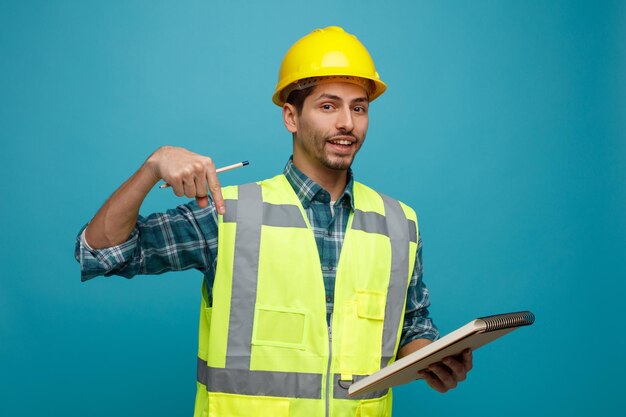 Joyeux jeune ingénieur masculin portant un casque de sécurité et un uniforme tenant un crayon et un bloc-notes regardant la caméra pointant vers le bas isolé sur fond bleu