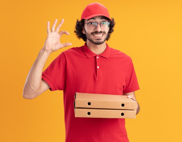 Joyeux jeune livreur caucasien en uniforme rouge et casquette portant des lunettes tenant des paquets de pizza faisant un geste de petite quantité isolé sur un mur orange