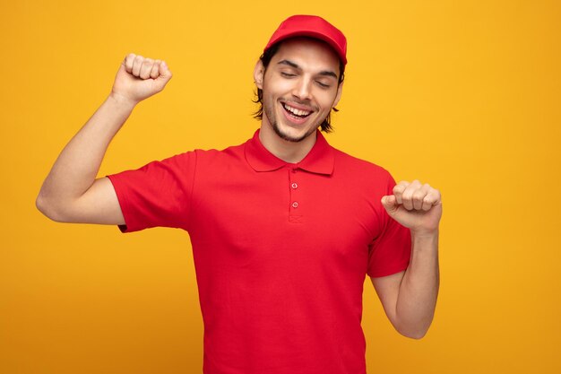 joyeux jeune livreur portant l'uniforme et la casquette gardant les poings dans l'air avec les yeux fermés isolé sur fond jaune