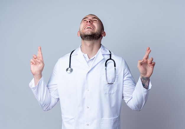 Joyeux jeune médecin de sexe masculin portant une robe médicale et un stéthoscope autour de son cou faisant le geste des doigts croisés avec les yeux fermés isolé sur blanc