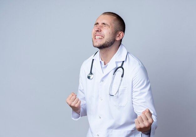 Joyeux jeune médecin de sexe masculin portant une robe médicale et un stéthoscope autour de son cou serrant les poings avec les yeux fermés isolé sur blanc