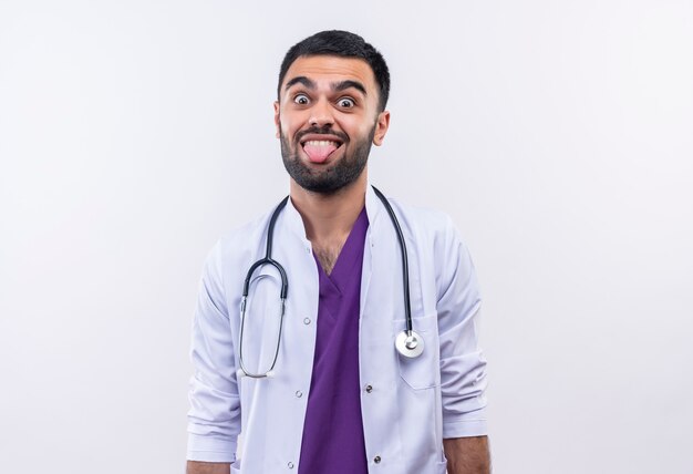 Joyeux jeune médecin de sexe masculin portant une robe médicale stéthoscope montrant la langue sur un mur blanc isolé