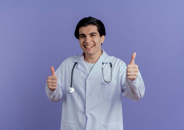 Joyeux jeune médecin de sexe masculin portant une robe médicale et un stéthoscope montrant les pouces vers le haut isolé sur un mur violet avec copie espace