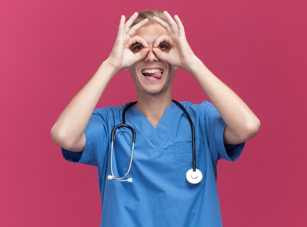 Joyeux jeune médecin de sexe masculin portant l'uniforme de médecin avec stéthoscope montrant la langue et le geste de regard isolé sur mur rose