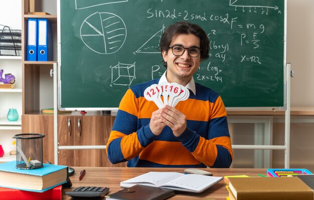 Joyeux jeune professeur de géométrie caucasien portant des lunettes assis au bureau avec des fournitures scolaires en classe montrant le nombre de fans regardant à l'avant
