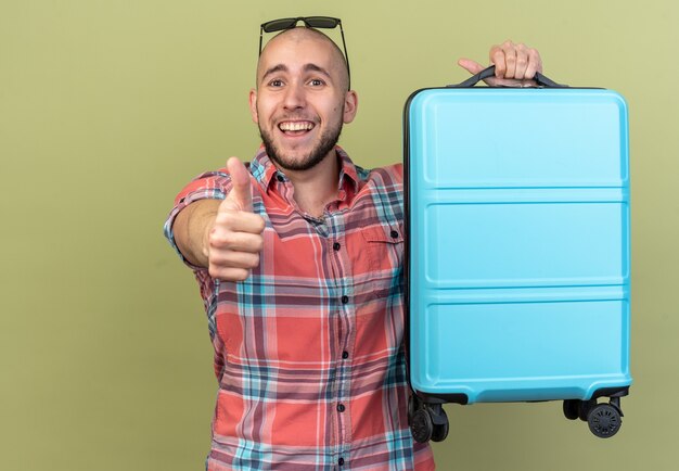 joyeux jeune voyageur homme tenant une valise et le pouce levé isolé sur un mur vert olive avec espace pour copie