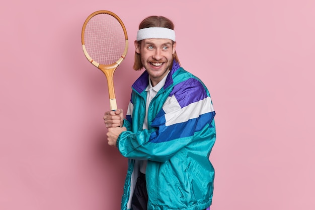 Joyeux joueur barbu tient la raquette de tennis aime le jeu actif sur le court vêtu de vêtements de sport a une expression heureuse regarde à distance.