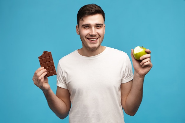 Joyeux joyeux jeune homme aux cheveux noirs regardant la caméra avec un large sourire excité tenant une pomme verte à moitié mordue