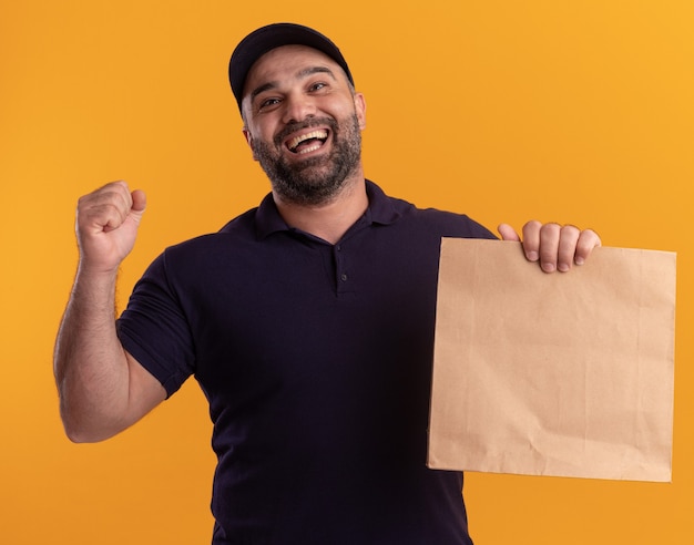 Joyeux livreur d'âge moyen en uniforme et cap holding paper food package montrant oui geste isolé sur mur jaune