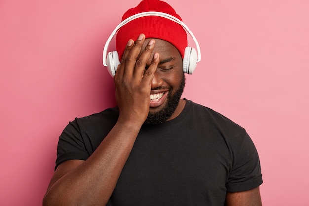 Joyeux mec heureux au chapeau rouge, ferme les yeux, sourit largement, choisit une chanson pour se détendre porte un casque stéréo, isolé sur un mur rose, passe du temps libre à écouter de la musique.