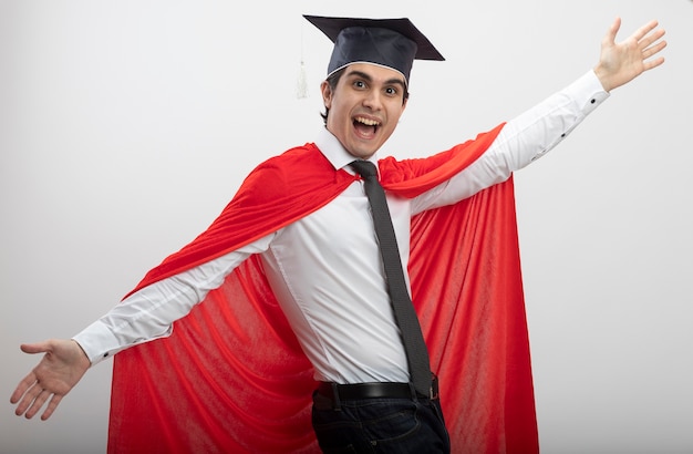 Joyeux mec jeune super-héros regardant la caméra portant une cravate et un chapeau diplômé répandant les mains isolé sur fond blanc