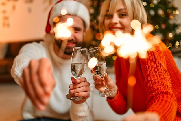 Joyeux noel et bonne année! un couple d'amoureux portant des chapeaux de père noël et des bois de renne assis sur le sol près d'un arbre de noël avec des cierges magiques et des verres de champagne, attendant des vacances.