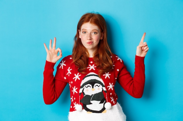 Joyeux Noël. Joyeuse rousse en pull de Noël, pointant le doigt dans le coin supérieur droit, montrant la promo du nouvel an et d'accord avec l'approbation, louez le produit.