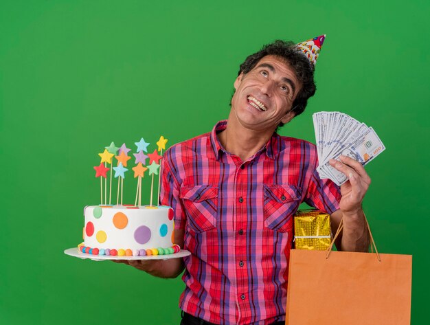 Joyeux parti caucasien d'âge moyen man wearing birthday cap holding birthday cake papier sac cadeau pack et de l'argent jusqu'à isolé sur fond vert