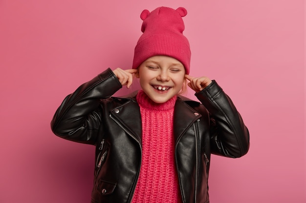 Joyeux petit enfant d'âge préscolaire couvre les oreilles, garde les index dans les trous d'oreille, évite d'entendre de la musique forte, a une expression heureuse, porte un chapeau rose avec des oreilles et une veste en cuir, ne veut pas entendre de bruit