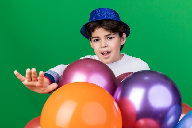 Joyeux petit garçon portant un chapeau de fête bleu debout derrière des ballons tenant la main isolée sur un mur vert