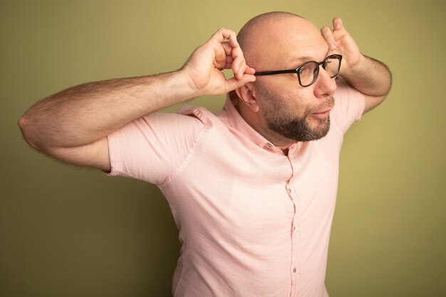 Joyeux regardant côté homme chauve d'âge moyen portant un t-shirt rose portant et tenant des lunettes isolé sur mur vert olive