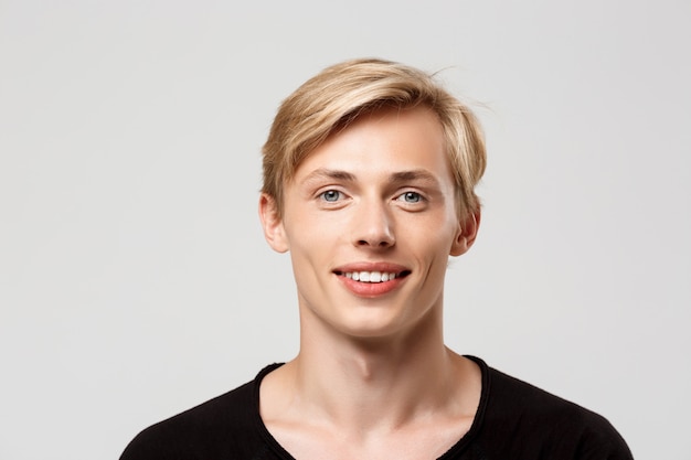 Joyeux sourire blond beau jeune homme portant un t-shirt noir sur le mur gris