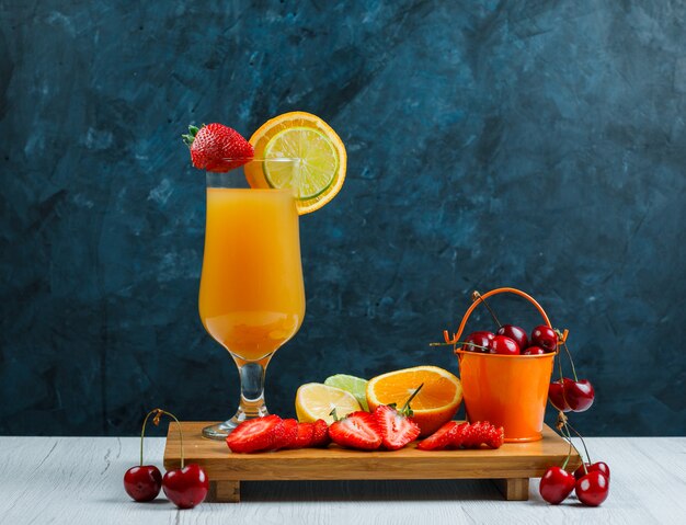 Jus d'orange dans un gobelet avec agrumes, fraise, cerise, planche à découper vue latérale sur fond bleu en bois et grungy