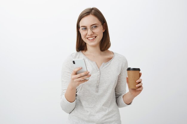 Juste une seconde besoin de réponse. Charmante belle femme européenne insouciante dans des verres tenant une tasse de papier avec une boisson de café appréciant parler avec le patron et taper un message dans le smartphone sur le mur gris