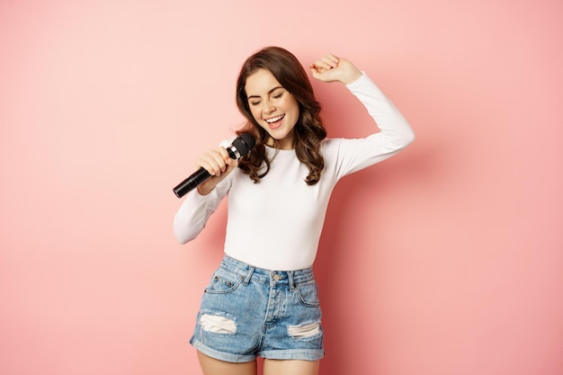 Photo gratuite karaoké. heureuse fille souriante chantant dans le microphone, dansant et s'amusant, debout sur fond rose