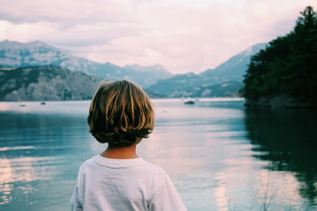 Kid aux cheveux blonds regardant la mer avec des montagnes au loin tourné par derrière