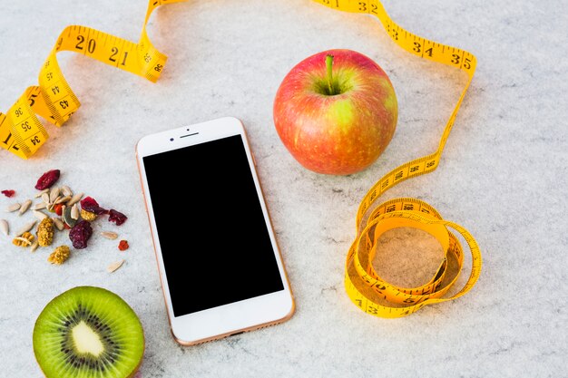 Kiwi réduit de moitié; fruits secs; Pomme; ruban à mesurer et smartphone sur fond gris texturé