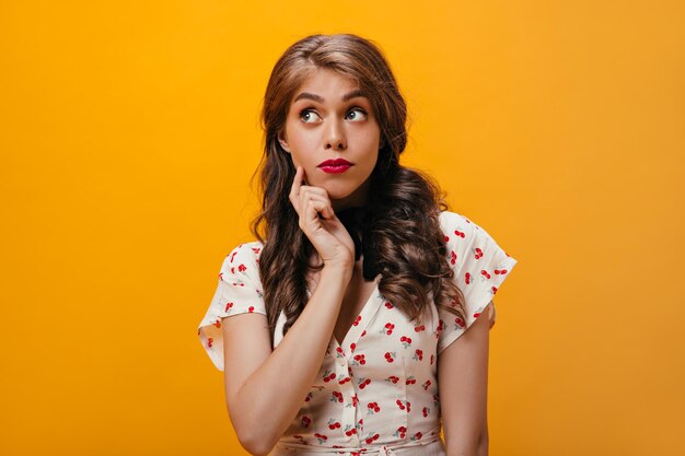 Lady pose pensivement sur fond orange Pensive curly young woman with pink lips in trendy outfit looking up on isolated background