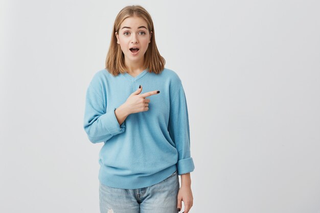 Le langage du corps. Fille choquée, excitée et surprise avec les cheveux blonds raides et la bouche ouverte, les yeux embêtés, pointant avec l'index à l'espace de copie pour votre texte promotionnel ou votre publicité