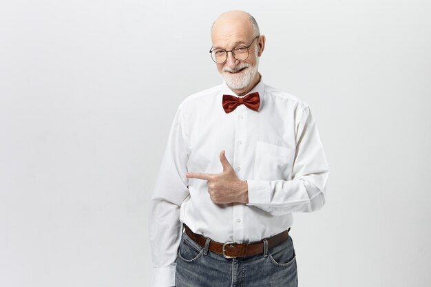 Le langage du corps. Heureux homme âgé positif en jeans décontractés, lunettes, chemise et nœud papillon rouge souriant, pointant l'index loin, indiquant un mur de fond vierge pour votre contenu publicitaire