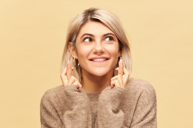 Le langage du corps. Portrait de l'adorable jeune femme superstitieuse mignonne aux cheveux blonds et anneau de nez croisant les doigts pour la bonne chance, souhaitant gagner à la loterie, ses yeux pleins d'espoir et d'impatience