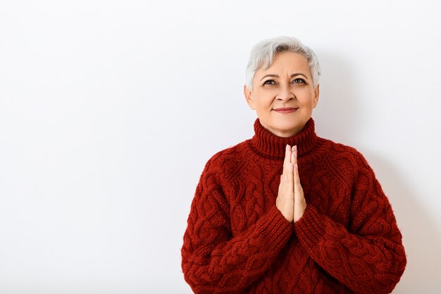 Le langage du corps. Tir horizontal de la charmante belle femme aux cheveux gris à la retraite posant au mur dans un pull tricoté confortable, serrant les mains ensemble et souriant largement, excité par de bonnes nouvelles