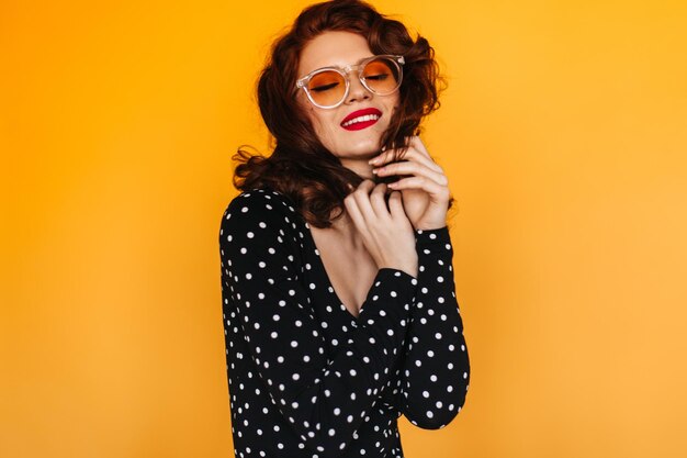 Laughing woman in polkadot shirt standing on yellow background Studio shot of ginger girl in sunglasses