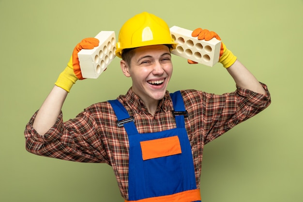Photo gratuite laughing young male builder en uniforme avec des gants tenant des briques autour de la tête