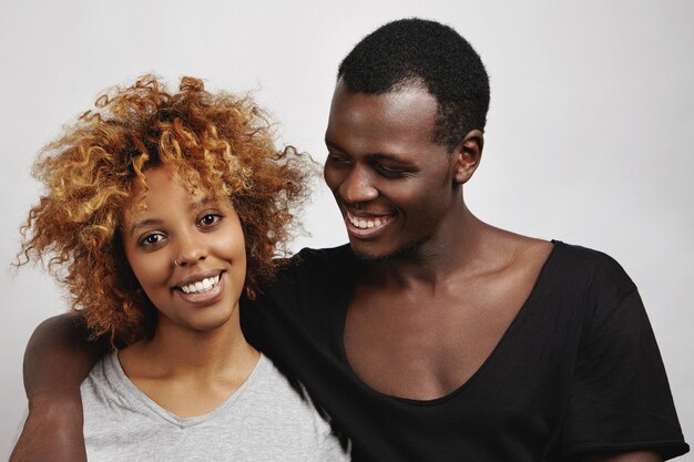 Élégant couple africain posant: heureux bel homme en t-shirt noir serrant sa belle petite amie avec une coiffure afro et un anneau dans le nez, la regardant avec un sourire affectueux.