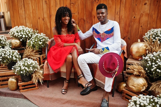 Élégant couple afro france à la mode posé ensemble au jour de l'automne Modèles africains noirs amoureux assis contre une décoration en bois avec des fleurs et des citrouilles