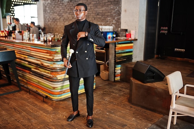 Élégant gentleman afro-américain dans une élégante veste noire et des lunettes tenant une canne rétro comme un ballon de canne ou une canne basculante avec une poignée en boule de diamant doré Riche homme afro à la mode