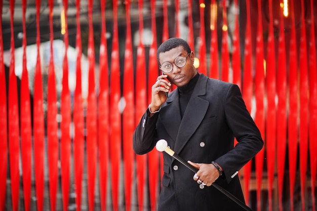 Élégant gentleman afro-américain dans une élégante veste noire tenant un bâton de marche rétro comme flacon de canne ou canne à bascule Riche homme afro à la mode contre le mur rouge parlant au téléphone mobile
