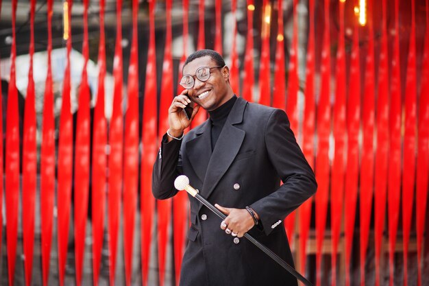 Élégant gentleman afro-américain dans une élégante veste noire tenant un bâton de marche rétro comme flacon de canne ou canne à bascule Riche homme afro à la mode contre le mur rouge parlant au téléphone mobile