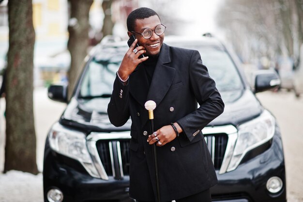 Élégant gentleman afro-américain dans une élégante veste noire tenant un bâton de marche rétro comme flacon de canne ou canne à bascule Riche homme afro à la mode contre une voiture suv d'affaires parlant sur un téléphone mobile