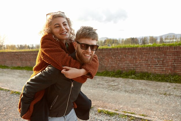 Élégant homme aux cheveux rouges mal rasé dans des tons rectangulaires portant sa jolie petite amie à la mode sur son dos, démontrant l'amour, les soins et l'affection. Heureux jeune couple à la mode se détendre à l'extérieur