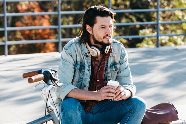 Élégant jeune homme assis dans le parc, tenant une tasse de café à emporter en mains