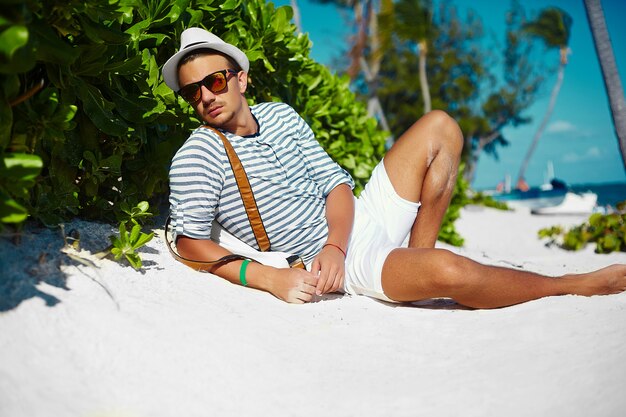 Élégant jeune homme modèle masculin allongé sur le sable de la plage portant un chapeau d'été hipster