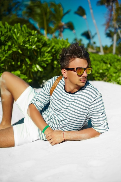 Élégant jeune homme modèle masculin allongé sur le sable de la plage portant un chapeau d'été hipster