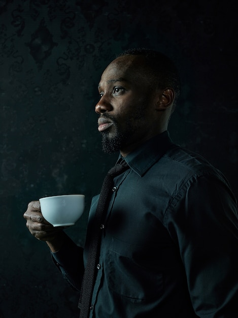 Élégant jeune homme noir africain avec une tasse blanche de café posant sur un studio sombre.