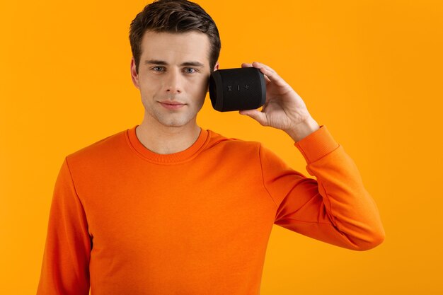 Élégant jeune homme souriant en pull orange tenant haut-parleur sans fil heureux d'écouter de la musique s'amuser sur orange