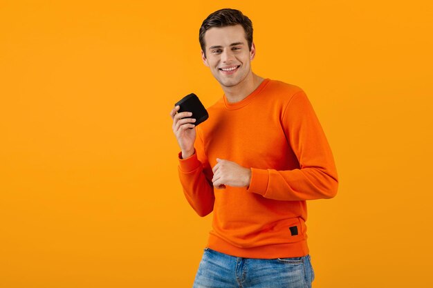 Élégant jeune homme souriant en pull orange tenant un haut-parleur sans fil heureux d'écouter de la musique s'amuser style coloré bonne humeur isolé sur fond jaune